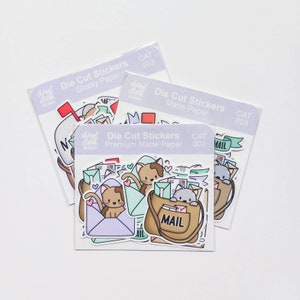 May include: Three packs of die-cut stickers featuring cute cats and mailboxes. The stickers are available in glossy and matte paper. The text on the sticker packs reads "Die Cut Stickers", "Glossy Paper", "Matte Paper", "Premium Matte Paper", "Cheryl Clark Designs", and "CAT 003".