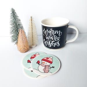 Peut inclure: Une tasse en céramique noire avec un lettrage blanc qui dit "Warm Winter Wishes". La tasse est posée sur trois dessous-de-verre ronds avec un motif de bonhomme de neige. Les dessous-de-verre sont bleu clair avec des flocons de neige blancs et un bonhomme de neige portant un chapeau et une écharpe roses.