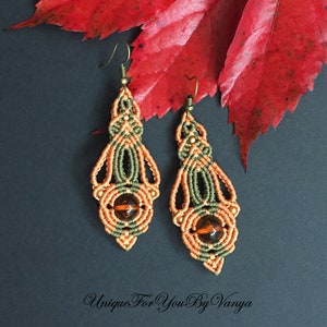 May include: A pair of dangle earrings with a green, orange, and gold macrame design. Each earring features a round amber-colored bead in the center.