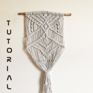May include: A white macrame wall hanging with a geometric pattern and a wooden dowel at the top. The hanging has a long tassel at the bottom. The text "TUTORIAL PDF" is written in black on a white background.