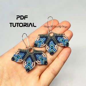 以下が含まれることがあります： 青、緑、グレーの色合いで幾何学模様が施された、手作りのマクラメイヤリングのペア。イヤリングは小さな丸いビーズで飾られており、シルバーのフックが付いています。