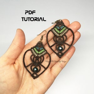 Peut inclure: Une paire de boucles d'oreilles en macramé marron et vert en forme de cœur avec une perle de cristal vert foncé au centre. Les boucles d'oreilles sont suspendues à des crochets en bronze. Le texte "PDF TUTORIAL" est en haut de l'image.