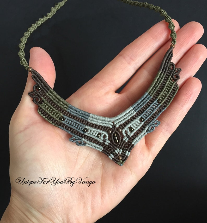May include: A brown, grey and green macrame necklace with small brown beads. The necklace is in the shape of a V and has a delicate, intricate design.