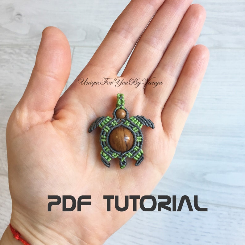 May include: A green and gray macrame turtle pendant with a brown wood bead in the center. The pendant is held in a person's hand. The text "PDF TUTORIAL" is printed below the pendant.