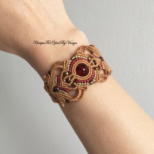 May include: A brown and red macrame bracelet with a red gemstone in the center. The bracelet is decorated with gold beads and black beads. "Unique For You By Vanya" is written in black text on the bracelet.