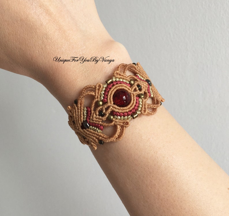 May include: A brown and red macrame bracelet with a red gemstone in the center. The bracelet is decorated with gold beads and black beads. "Unique For You By Vanya" is written in black text on the bracelet.