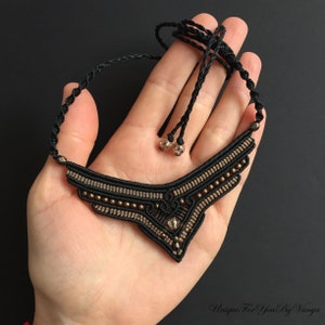 May include: A black macrame necklace with brown beads and a geometric design. The necklace is held in a hand.