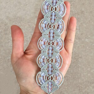 Puede incluir: Un brazalete de macram&eacute; gris con cuentas de color azul claro. El brazalete est&aacute; hecho con una t&eacute;cnica de anudado y tiene un cierre plateado.