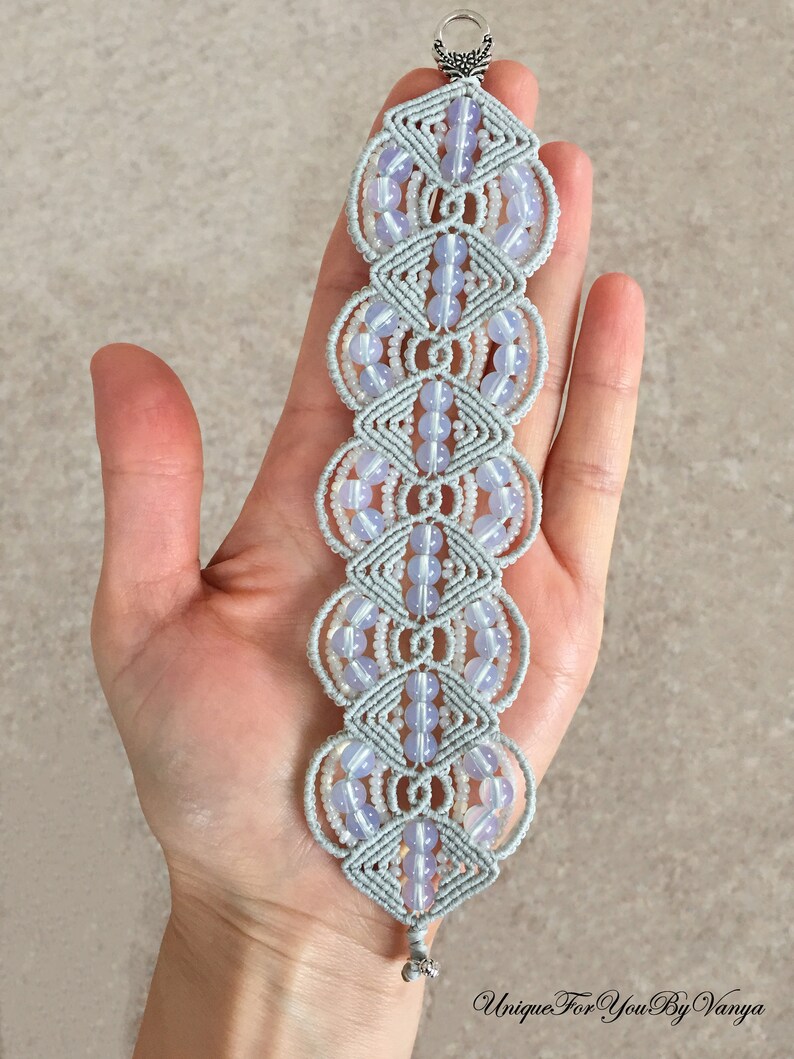 Puede incluir: Un brazalete de macram&eacute; gris con cuentas de color azul claro. El brazalete est&aacute; hecho con una t&eacute;cnica de anudado y tiene un cierre plateado.