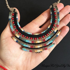 K&ouml;nnte beinhalten: Eine bunte, mehrlagige Makramee-Halskette mit roten, schwarzen, beigen, blauen und wei&szlig;en Perlen. Die Halskette hat die Form eines Halbmondes und wird in einer Hand gehalten.