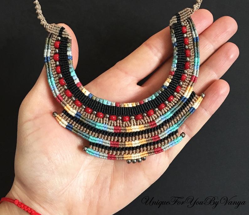 K&ouml;nnte beinhalten: Eine bunte, mehrlagige Makramee-Halskette mit roten, schwarzen, beigen, blauen und wei&szlig;en Perlen. Die Halskette hat die Form eines Halbmondes und wird in einer Hand gehalten.