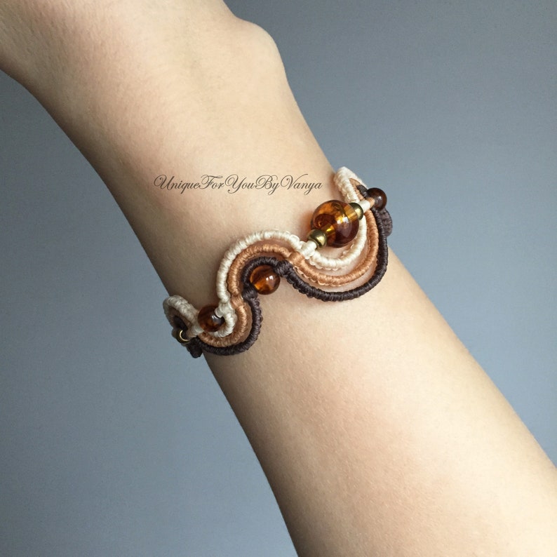 K&ouml;nnte beinhalten: Ein braunes, beiges und wei&szlig;es Makramee-Armband mit bernsteinfarbenen Glasperlen. Das Armband ist in einem wellenf&ouml;rmigen Muster gebunden.