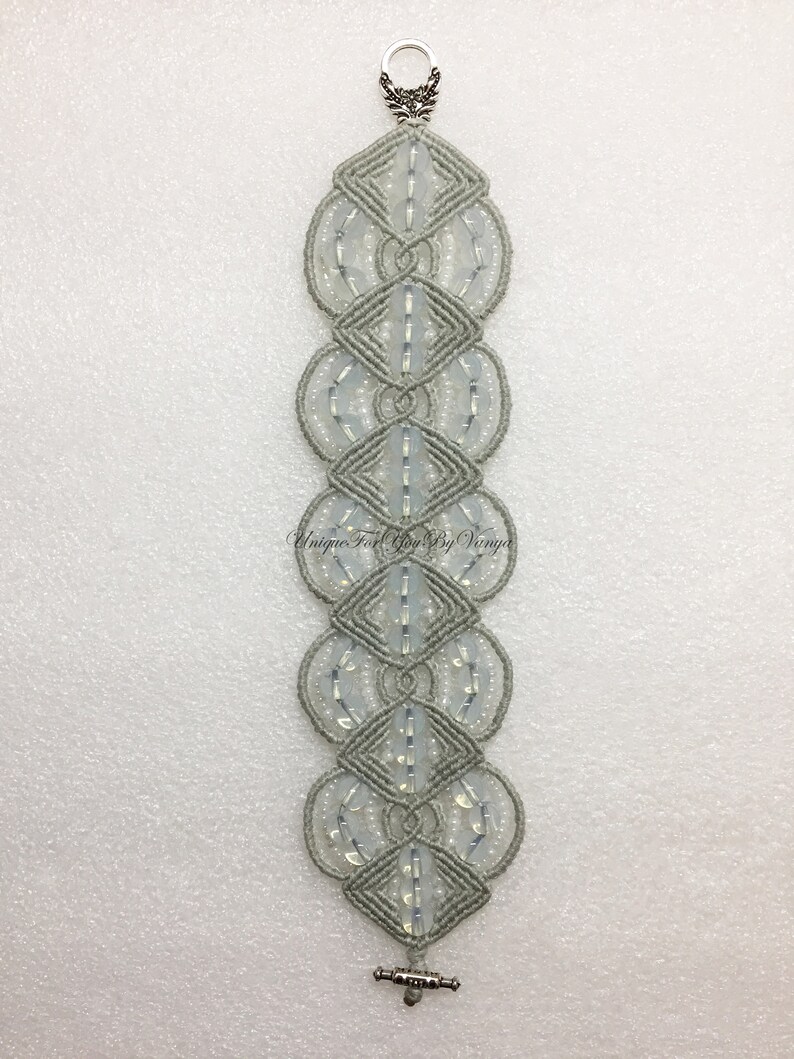 Puede incluir: Un brazalete de macram&eacute; gris con cuentas blancas y cierres plateados. "Unique For You By Vinya" est&aacute; escrito en el brazalete.
