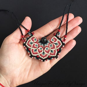 Może przedstawiać: Ręcznie robiony naszyjnik z makramą z czarnym kamieniem jako zawieszką. Naszyjnik wykonany jest z czerwonej, białej i niebieskiej sznur&oacute;wki i ma geometryczny wz&oacute;r.