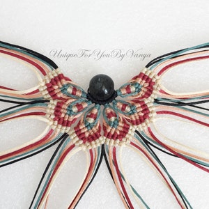 Może przedstawiać: Zbliżenie na projekt makramy z czarną koralikiem w środku. Projekt wykonany jest z czerwonych, białych i turkusowych sznurk&oacute;w.