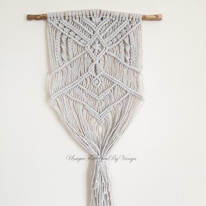 May include: A white macrame wall hanging with a geometric pattern and a long tassel. The hanging is attached to a wooden dowel.