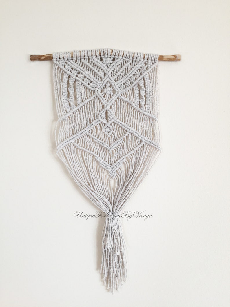 May include: A white macrame wall hanging with a geometric pattern and a long tassel. The hanging is attached to a wooden dowel.