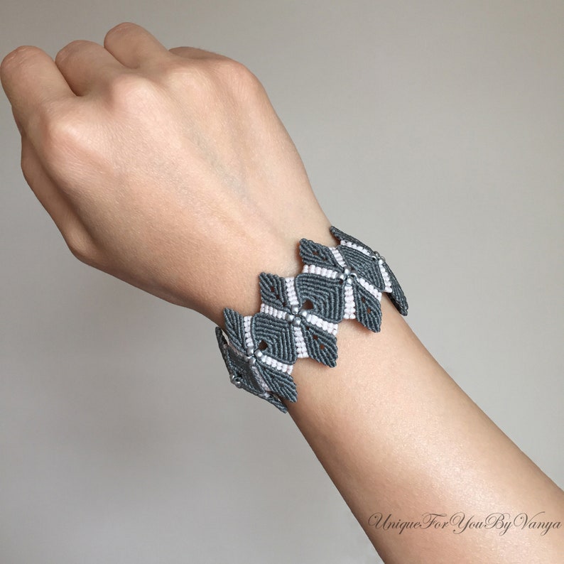 May include: Grey and white macrame bracelet with a geometric pattern. The bracelet is made using a knotting technique.