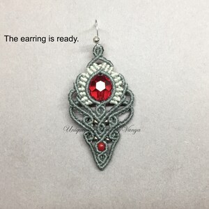 Pu&ograve; includere: Un orecchino in macram&eacute; grigio e bianco con un cristallo rosso al centro. L'orecchino &egrave; appeso a un gancio argentato.