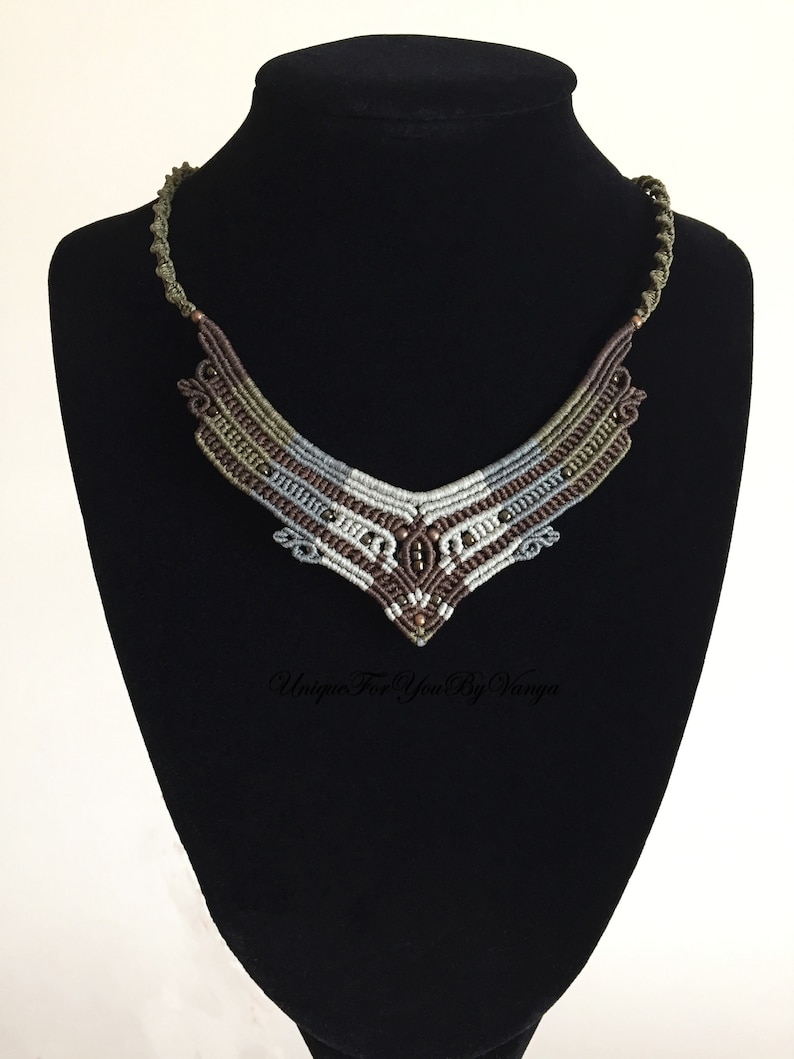 May include: A brown, grey and green macrame necklace with beads. The necklace is in the shape of a V and is displayed on a black mannequin.