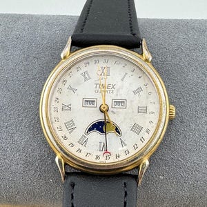 Peut inclure: Une montre Timex Quartz dorée avec un cadran blanc, des chiffres romains pour les heures et un indicateur de phase lunaire. La montre a un bracelet en cuir noir et affiche le jour et la date. Le cadran affiche également le jour de la semaine et le mois.