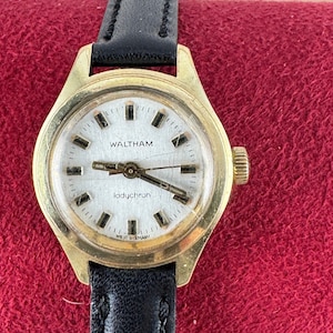 Op de afbeelding: Een goudkleurig polshorloge met een witte wijzerplaat en een zwart leren bandje. De wijzerplaat van het horloge heeft zwarte Romeinse cijfers als uurmarkeringen en de tekst "WALTHAM Ladychron WEST GERMANY".