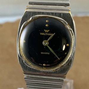 Puede incluir: Un reloj de pulsera de cuarzo Waltham plateado y dorado con una esfera negra y una pulsera de metal plateado.