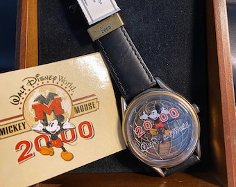 Könnte beinhalten: Eine Armbanduhr aus schwarzem Leder mit einem goldfarbenen Gehäuse und einem weißen Zifferblatt. Das Zifferblatt zeigt eine Mickey-Mouse-Illustration und den Text "Walt Disney World 2000". Die Uhr wird in einer Holzkiste mit einem Echtheitszertifikat geliefert.