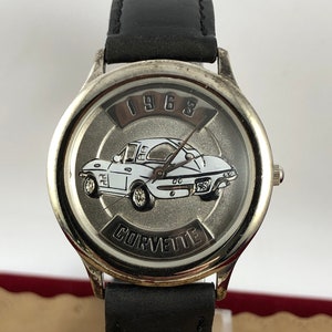 May include: A silver-toned wristwatch with a black leather band. The watch face features a white 1965 Corvette with the text "1965" and "Corvette" on the face.