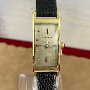 May include: A vintage gold-plated rectangular watch with the brand name "VALDEAN" on the face. The watch has a light-coloured dial, gold hour markers, and black hands. It features a black leather strap.