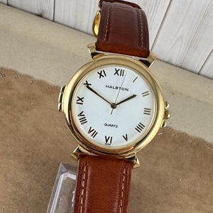 May include: A gold-tone watch with a white face and Roman numeral hour markers. The watch has a brown leather strap and says "Halbton Quartz" on the face.