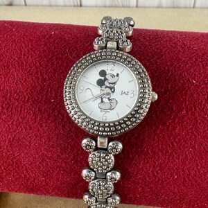 Puede incluir: Un reloj Mickey Mouse plateado con una esfera blanca y una imagen de Mickey Mouse negra. El reloj tiene una correa plateada con eslabones con forma de cabeza de Mickey Mouse. La esfera del reloj tiene n&uacute;meros y manecillas negros, y el texto "JAZ" y "&copy;Disney" impreso en &eacute;l.