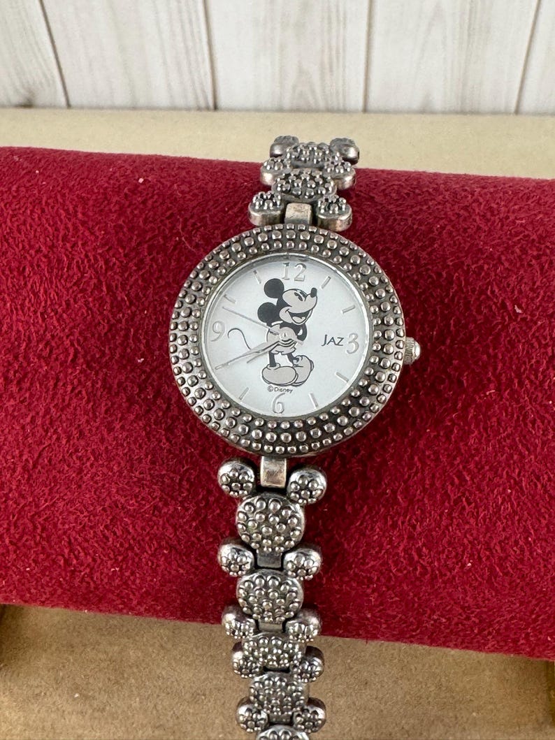 Puede incluir: Un reloj Mickey Mouse plateado con una esfera blanca y una imagen de Mickey Mouse negra. El reloj tiene una correa plateada con eslabones con forma de cabeza de Mickey Mouse. La esfera del reloj tiene n&uacute;meros y manecillas negros, y el texto "JAZ" y "&copy;Disney" impreso en &eacute;l.