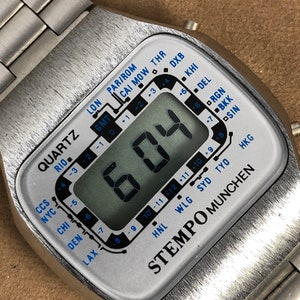 Op de afbeelding: Een zilveren digitale horloge met een witte wijzerplaat en een zwart digitaal display. De wijzerplaat van het horloge heeft een tijdzoneweergave met de tijd in verschillende steden over de hele wereld. De wijzerplaat van het horloge heeft ook de woorden "STEMPO MUNCHEN" erop gedrukt.