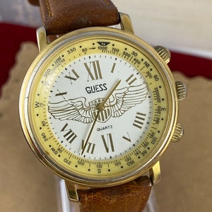 May include: A gold-toned Guess watch with a white face and Roman numeral hour markers. The watch has a brown leather strap and features a compass and tachymeter scale around the outer edge of the face. The words "GUESS" and "QUARTZ" are printed on the face.