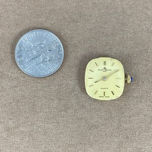 May include: A square-shaped, gold-toned watch with a white face and black hands. The watch face has the text "BAUME & MERCIER" and "QUARTZ" printed on it. The watch is made in Switzerland.