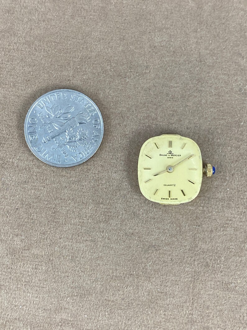 May include: A square-shaped, gold-toned watch with a white face and black hands. The watch face has the text "BAUME & MERCIER" and "QUARTZ" printed on it. The watch is made in Switzerland.