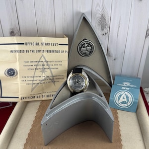 May include: A Star Trek watch with a black leather strap, presented in a silver case shaped like the Starfleet emblem. The watch face has a detailed design. A certificate of authenticity is also included.