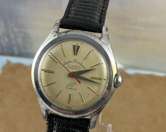 May include: A silver Andre Boucher 17 Jewels Automatic watch with a black leather band. The watch face is white with black Roman numeral hour markers and black hands. The watch has a red second hand and a date window at the 3 o'clock position. The watch also has a SS Incabloc inscription.