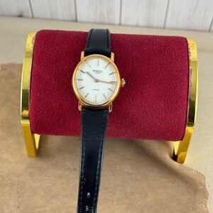 K&ouml;nnte beinhalten: Eine goldfarbene Armbanduhr mit wei&szlig;em Zifferblatt und schwarzem Lederarmband. Das Zifferblatt hat schwarze r&ouml;mische Ziffern als Stundenmarkierungen und ein Breil-Logo.