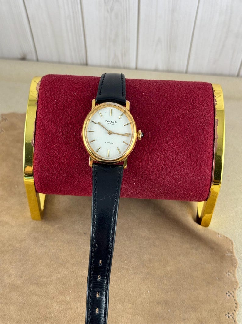 K&ouml;nnte beinhalten: Eine goldfarbene Armbanduhr mit wei&szlig;em Zifferblatt und schwarzem Lederarmband. Das Zifferblatt hat schwarze r&ouml;mische Ziffern als Stundenmarkierungen und ein Breil-Logo.