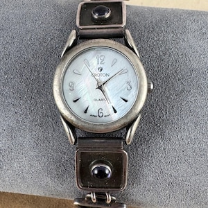 May include: Silver wristwatch with a white face and black Roman numeral hour markers. The watch has a silver band with black and red gemstones.