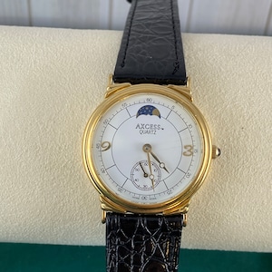 May include: A gold-tone watch with a white face and black leather band. The watch face has a moon phase indicator and the words "AXCESS QUARTZ" printed on it.