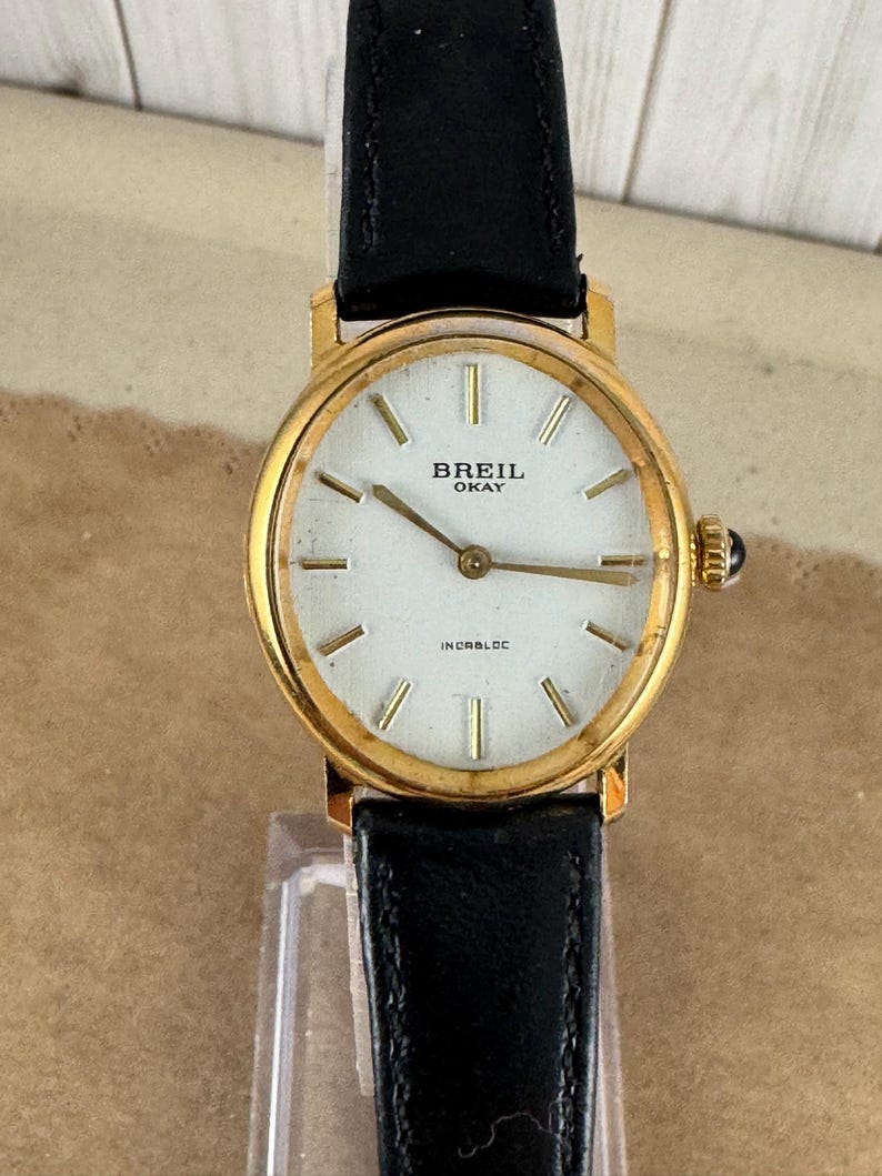 K&ouml;nnte beinhalten: Eine goldfarbene Breil Okay Armbanduhr mit wei&szlig;em Zifferblatt und schwarzem Lederband. Das Zifferblatt hat schwarze r&ouml;mische Ziffern als Stundenmarkierungen und einen schwarzen Sekundenzeiger. Auf dem Zifferblatt sind au&szlig;erdem die Texte "Breil Okay" und "Incabloc" aufgedruckt.
