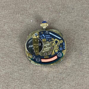 Baume and Mercier Quartz Watch Movement Swiss Made 6 Jewels BM5501 Sapphire Crown A gold-colored watch movement with a blue jewel on the winding stem. The movement has a gold dial with black markings and the text 'VB SWISS SIX 6 JEWELS' and '100 975 001'. The movement also has a silver-colored rotor with the text 'JOZA H1.4'.