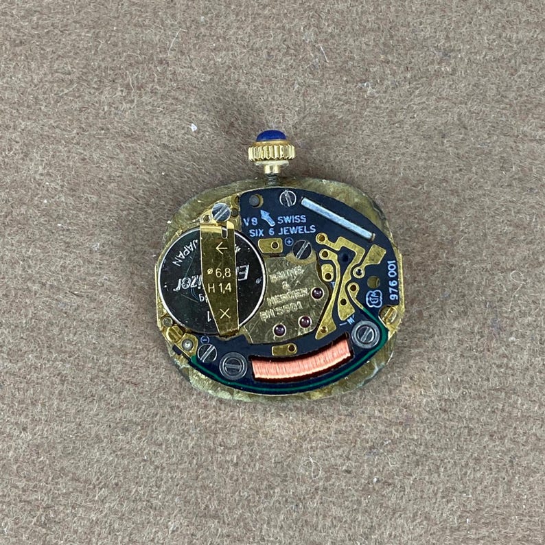 Baume and Mercier Quartz Watch Movement Swiss Made 6 Jewels BM5501 Sapphire Crown A gold-colored watch movement with a blue jewel on the winding stem. The movement has a gold dial with black markings and the text 'VB SWISS SIX 6 JEWELS' and '100 975 001'. The movement also has a silver-colored rotor with the text 'JOZA H1.4'.