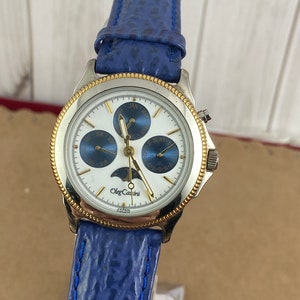 Könnte beinhalten: Eine silberne und goldfarbene Uhr mit einem blauen Lederarmband. Das Zifferblatt der Uhr ist weiß mit goldenen Akzenten und zeigt Tag, Datum und Mondphase an. Die Uhr ist mit "Oleg Cassini" und "Japan Movt." beschriftet.
