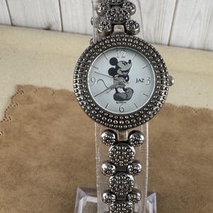 Puede incluir: Un reloj Mickey Mouse plateado con una banda plateada. La banda est&aacute; hecha de eslabones con forma de cabeza de Mickey Mouse. La esfera del reloj es blanca con im&aacute;genes de Mickey Mouse en negro y el texto "JAZ 3".