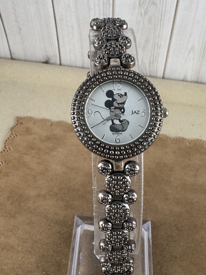 Puede incluir: Un reloj Mickey Mouse plateado con una banda plateada. La banda est&aacute; hecha de eslabones con forma de cabeza de Mickey Mouse. La esfera del reloj es blanca con im&aacute;genes de Mickey Mouse en negro y el texto "JAZ 3".