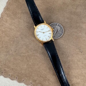 K&ouml;nnte beinhalten: Eine goldfarbene Breil-Uhr mit wei&szlig;em Zifferblatt und schwarzem Lederarmband. Die Uhr liegt auf einer braunen Wildlederoberfl&auml;che.